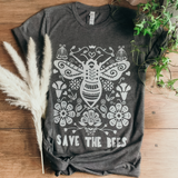On a rustic wooden table is a grey short-sleeved t-shirt with a white hand-drawn illustration. The illustration shows a big bumblebee surrounded by stylized flowers and a slogan at the bottom that says "save the bees".